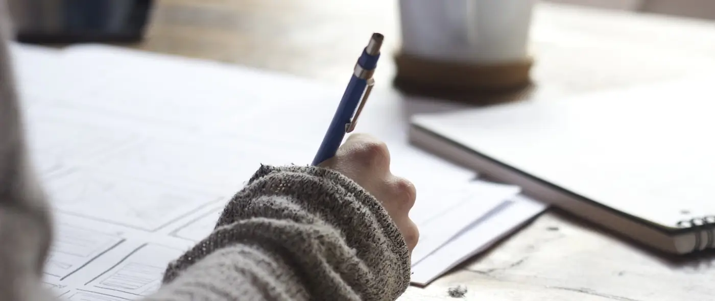 Person writing in a notebook at a wooden desk with a cup of coffee, reviewing notes