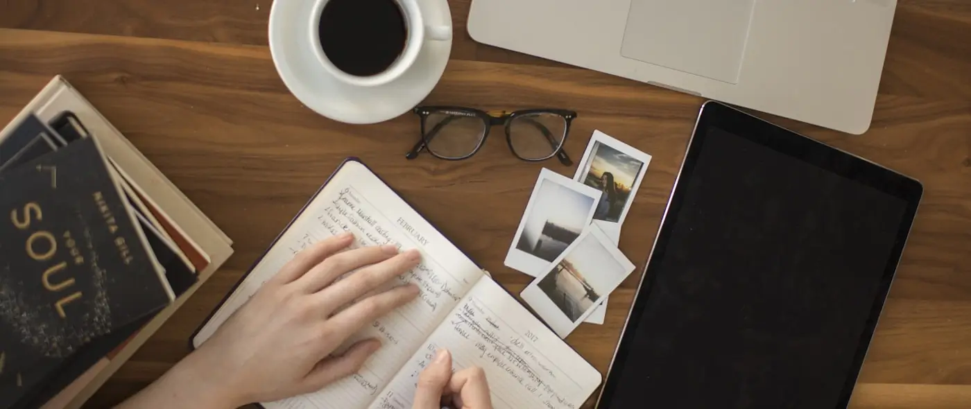 Person writing notes in a notebook beside an open laptop on a wooden desk